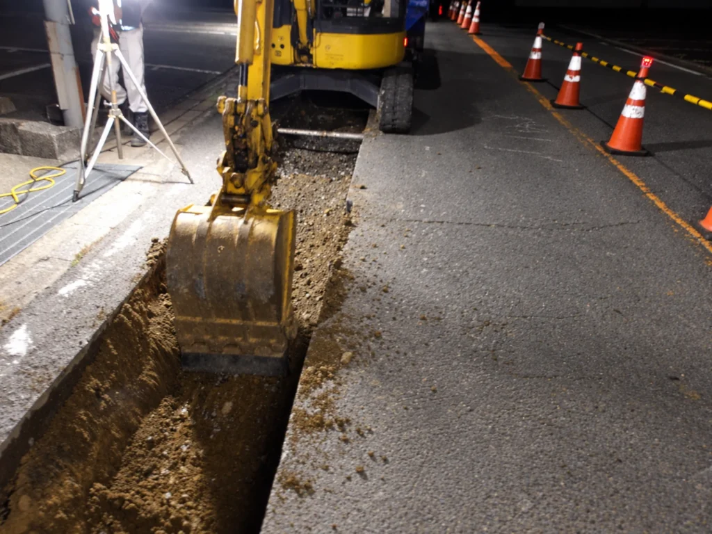 小榊線線路変更工事（管路工事）：夜間に道路の管路施工箇所をバックホウで掘削する作業