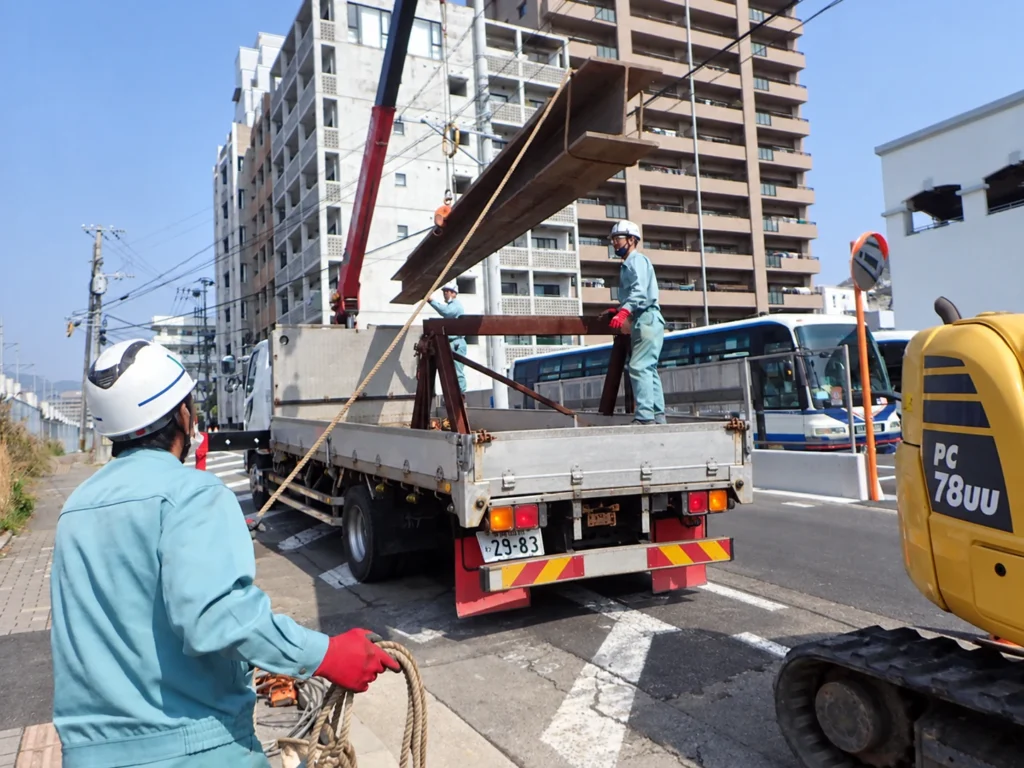 長崎駅周辺土地区画整理事業（2工区）電線共同溝工事：覆工板の撤去に伴い受け桁の鋼材を吊り上げて搬出する作業